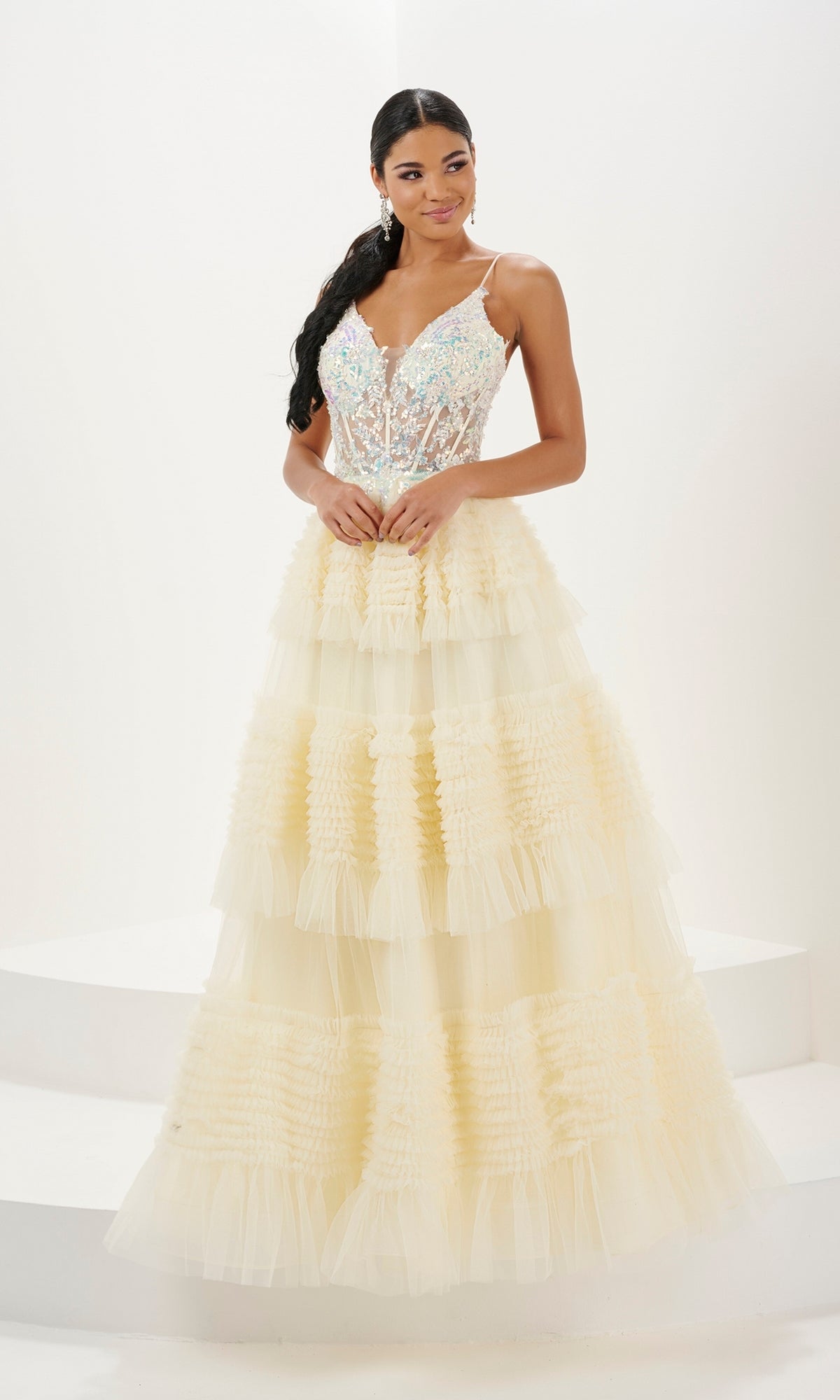 A woman stands on a white background wearing the Long Prom Dress 16054 by Tiffany—a sleeveless, tiered pale yellow A-line ball gown with an embellished V-neck and sheer bodice.